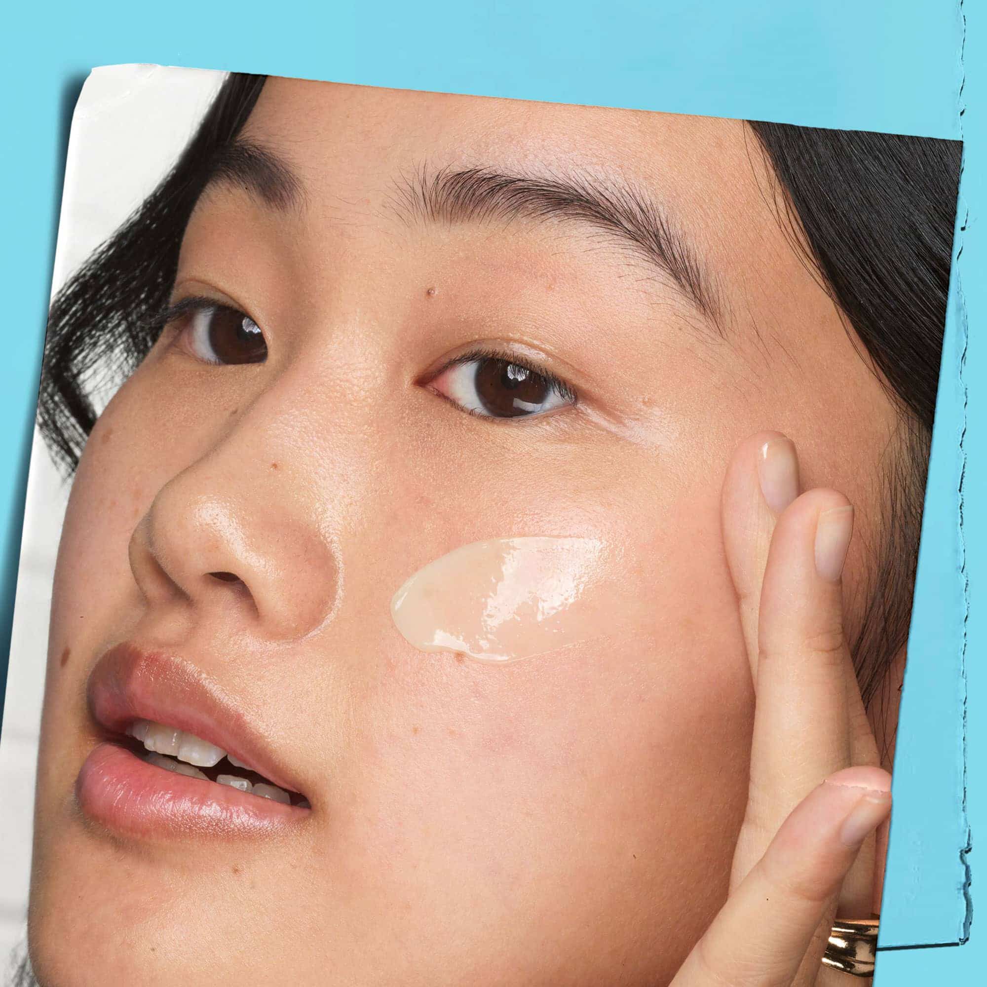 A graphic featuring a close-up of a person's face. A hand is gently touching the cheek, where a dab of light-colored product is visible. The image is set against a light blue background.
