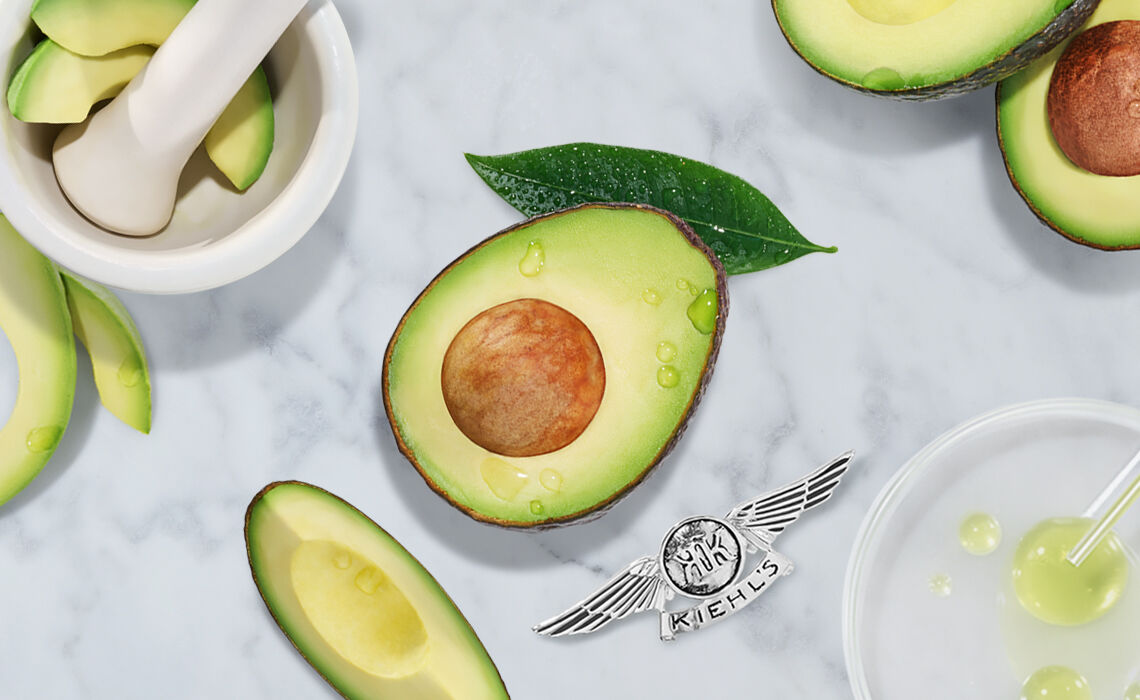 An arrangement of avocado slices, a whole avocado cut in half with a visible seed, and green leaves on a marble surface. A mortar and pestle and some liquid droplet elements are also present, along with the Kiehl's logo at the bottom.