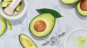 An arrangement of avocado slices, a whole avocado cut in half with a visible seed, and green leaves on a marble surface. A mortar and pestle and some liquid droplet elements are also present, along with the Kiehl's logo at the bottom.