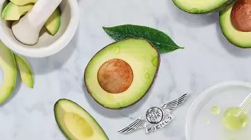 An arrangement of avocado slices, a whole avocado cut in half with a visible seed, and green leaves on a marble surface. A mortar and pestle and some liquid droplet elements are also present, along with the Kiehl's logo at the bottom.