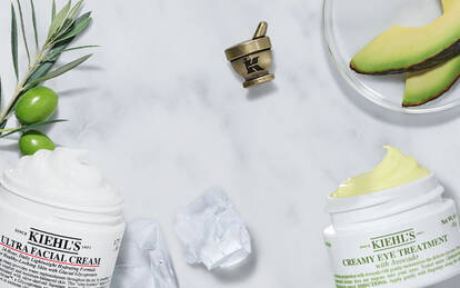 Visuel de la gamme Kiehl's presentant l'Ultra Facial Cream et le Creamy Eye Treatment with Avocado, disposes sur un fond de marbre blanc avec des tranches d'avocat frais, des olives vertes, des glacons et des gouttes de serum.