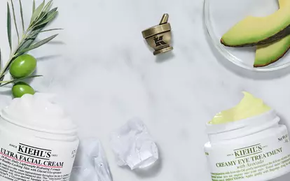 Visuel de la gamme Kiehl's presentant l'Ultra Facial Cream et le Creamy Eye Treatment with Avocado, disposes sur un fond de marbre blanc avec des tranches d'avocat frais, des olives vertes, des glacons et des gouttes de serum.