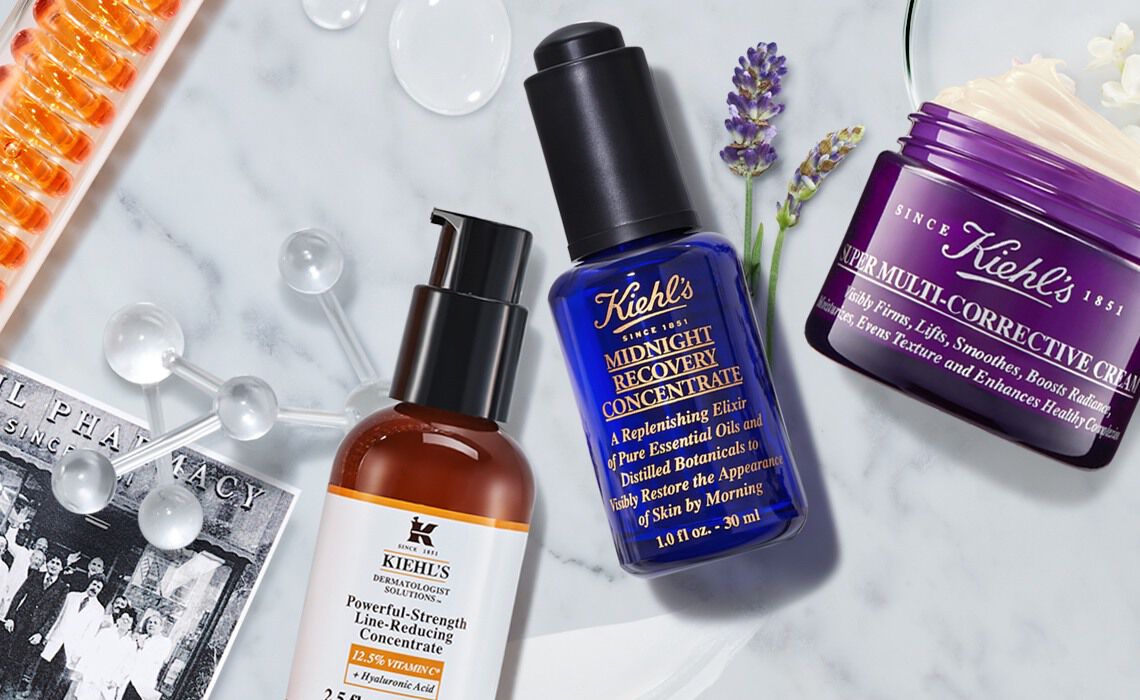 Image shows an arrangement of Kiehl's skincare products, including a serum and a cream, alongside ingredients like lavender and other skincare elements, set against a light background.
