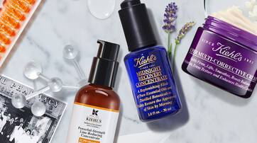 Image shows an arrangement of Kiehl's skincare products, including a serum and a cream, alongside ingredients like lavender and other skincare elements, set against a light background.