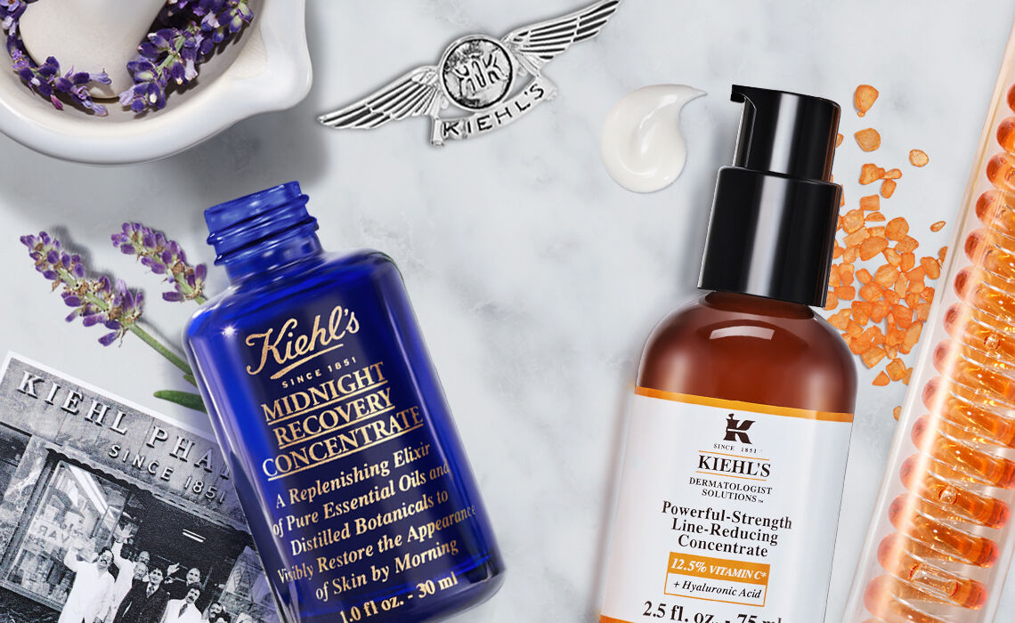 A flat lay of Kiehl's skincare products, featuring a blue Midnight Recovery Concentrate bottle, a brown Powerful-Strength Line-Reducing Concentrate, lavender flowers, a small Kiehl's label, and orange gel capsules, all arranged on a marble countertop.
