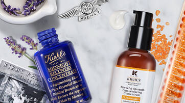 A flat lay of Kiehl's skincare products, featuring a blue Midnight Recovery Concentrate bottle, a brown Powerful-Strength Line-Reducing Concentrate, lavender flowers, a small Kiehl's label, and orange gel capsules, all arranged on a marble countertop.
