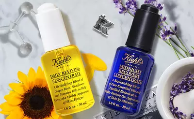 Alt text: Two Kiehl's skincare products, a yellow bottle labeled "Daily Reviving Concentrate" and a blue bottle labeled "Midnight Recovery Concentrate," placed on a light background with a sunflower and lavender.