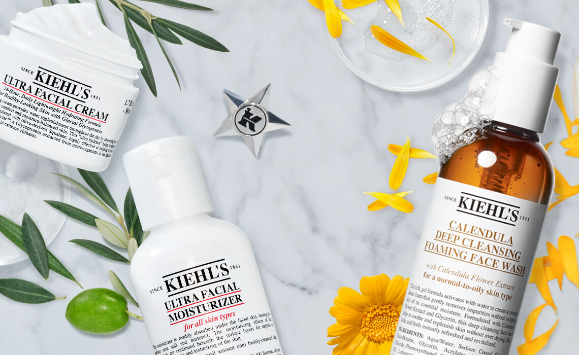 An arrangement of skincare products from Kiehl's on a marble surface, including Ultra Facial Cream, Ultra Facial Moisturizer, and Calendula Deep Cleansing Foaming Face Wash, surrounded by yellow flowers and green leaves.