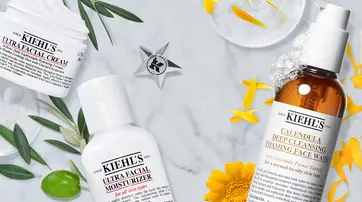 An arrangement of skincare products from Kiehl's on a marble surface, including Ultra Facial Cream, Ultra Facial Moisturizer, and Calendula Deep Cleansing Foaming Face Wash, surrounded by yellow flowers and green leaves.