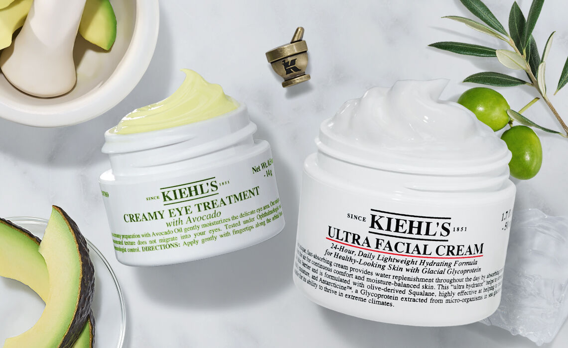 Two jars of Kiehl's skincare products are displayed on a marble surface. The jar on the left is labeled "Creamy Eye Treatment with Avocado," and the one on the right reads "Ultra Facial Cream." Surrounding the jars are avocado halves and a mortar and pestle.