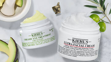 Two jars of Kiehl's skincare products are displayed on a marble surface. The jar on the left is labeled "Creamy Eye Treatment with Avocado," and the one on the right reads "Ultra Facial Cream." Surrounding the jars are avocado halves and a mortar and pestle.