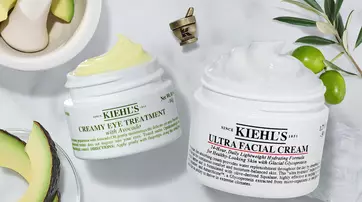 Two jars of Kiehl's skincare products are displayed on a marble surface. The jar on the left is labeled "Creamy Eye Treatment with Avocado," and the one on the right reads "Ultra Facial Cream." Surrounding the jars are avocado halves and a mortar and pestle.