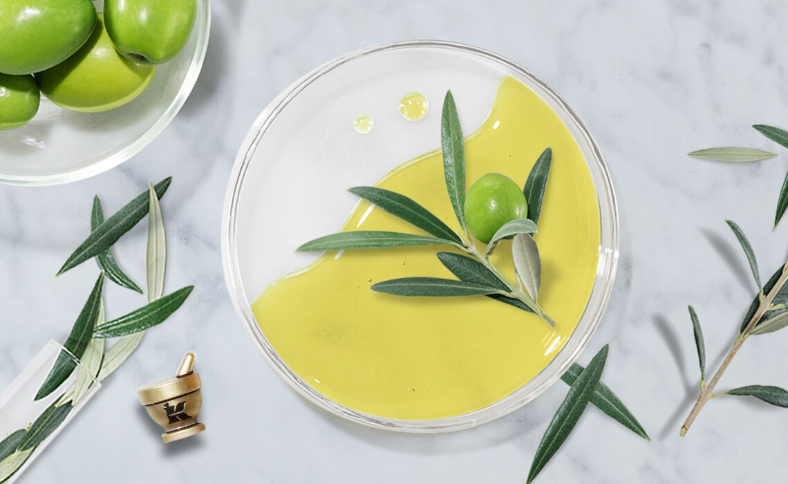 A clear dish filled with vibrant green olive oil, garnished with fresh olive leaves and an olive. A bowl of olives sits in the background, with scattered olive leaves and a small mortar and pestle nearby, all against a marble surface.