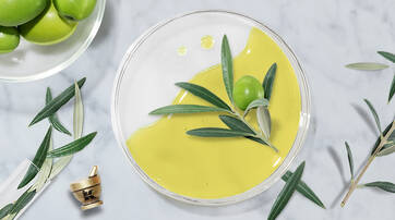 A clear dish filled with vibrant green olive oil, garnished with fresh olive leaves and an olive. A bowl of olives sits in the background, with scattered olive leaves and a small mortar and pestle nearby, all against a marble surface.