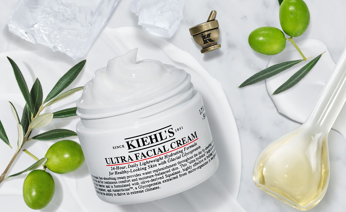 A jar of Kiehl's Ultra Facial Cream sits on a marble surface, surrounded by olive branches and olive oil, with a small decorative cup in the background. The cream is white and appears creamy in texture.