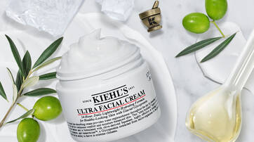 A jar of Kiehl's Ultra Facial Cream sits on a marble surface, surrounded by olive branches and olive oil, with a small decorative cup in the background. The cream is white and appears creamy in texture.