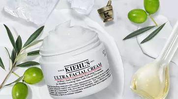 A jar of Kiehl's Ultra Facial Cream sits on a marble surface, surrounded by olive branches and olive oil, with a small decorative cup in the background. The cream is white and appears creamy in texture.
