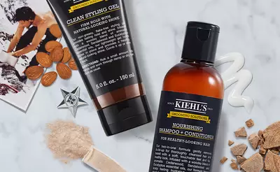 A flat lay of grooming products, featuring Kiehl's Clean Styling Gel and Nourishing Shampoo + Conditioner, surrounded by almond pieces, a star-shaped emblem, and scattered light-colored powders.