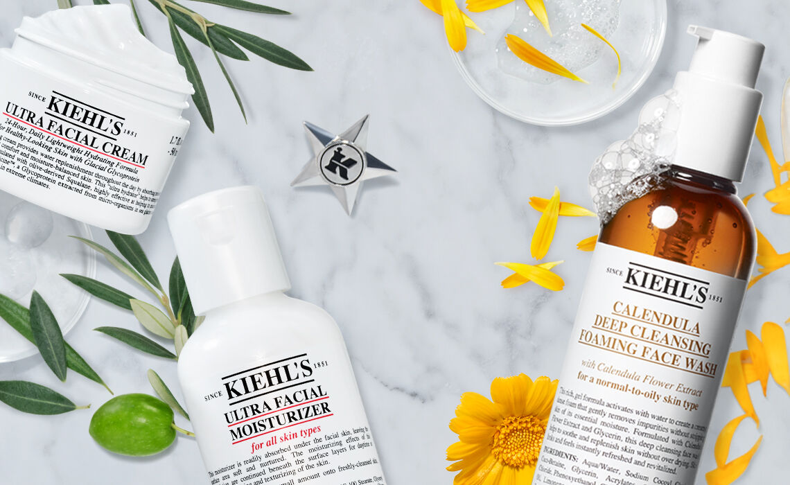 Flat lay of Kiehl's skincare products including Ultra Facial Cream, Ultra Facial Moisturizer, and Calendula Deep Cleansing Foaming Face Wash, surrounded by yellow flowers and greenery on a marble background.