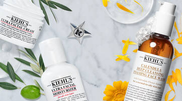 Flat lay of Kiehl's skincare products including Ultra Facial Cream, Ultra Facial Moisturizer, and Calendula Deep Cleansing Foaming Face Wash, surrounded by yellow flowers and greenery on a marble background.