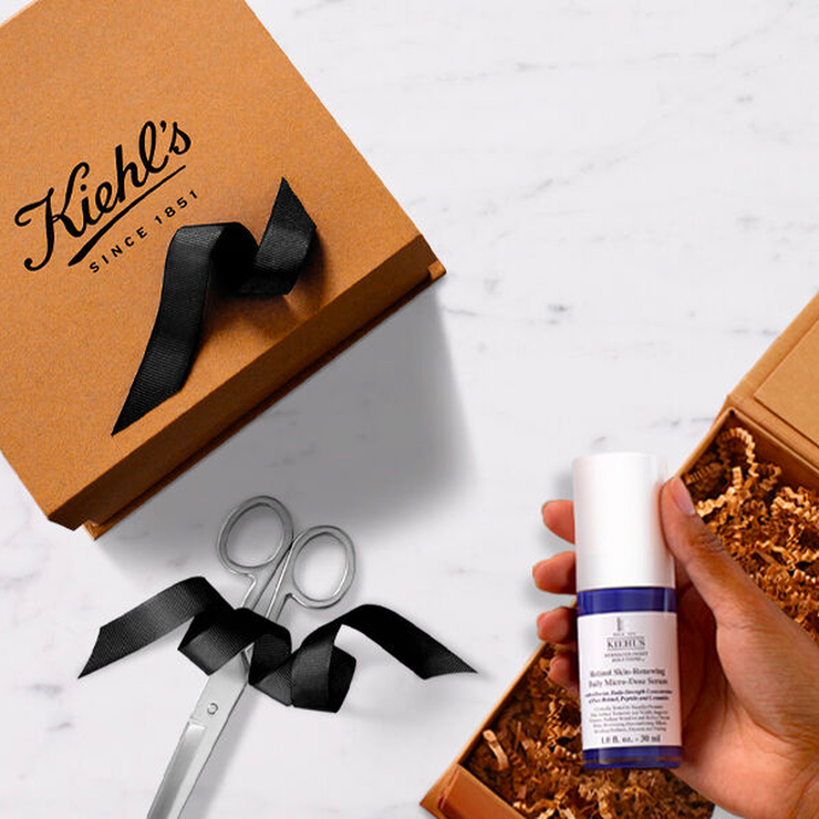 Coffret cadeau Kiehl’s en carton marron avec ruban noir, paire de ciseaux, et une main tenant un serum en flacon bleu et blanc, sur une table en marbre blanc.