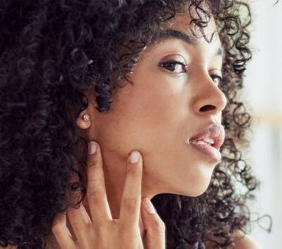 Femme a la peau eclatante et aux cheveux boucles, touchant delicatement son visage avec une expression douce.
