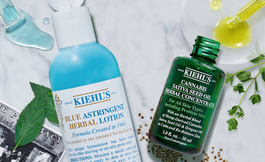 Deux produits de soin de la peau Kiehl's : un flacon de lotion astringente bleue et un flacon de concentre d'huile de graine de cannabis, entoures de feuilles et d'ingredients naturels.