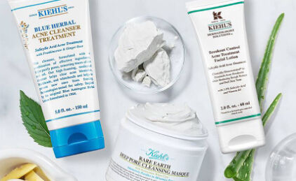 Produits de soins de la peau de la marque Kiehl's, comprenant un nettoyant pour l'acne, une lotion et un masque en creme, accompagnes de feuilles d'aloes et d'un bol de produit.