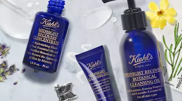 Trois produits de soins de la peau de Kiehl's : un flacon bleu de concentre de recuperation de minuit, un tube de creme pour les yeux et un flacon de nettoyant a l'huile botanique, entoures de gouttes d'huile et d'une fleur.